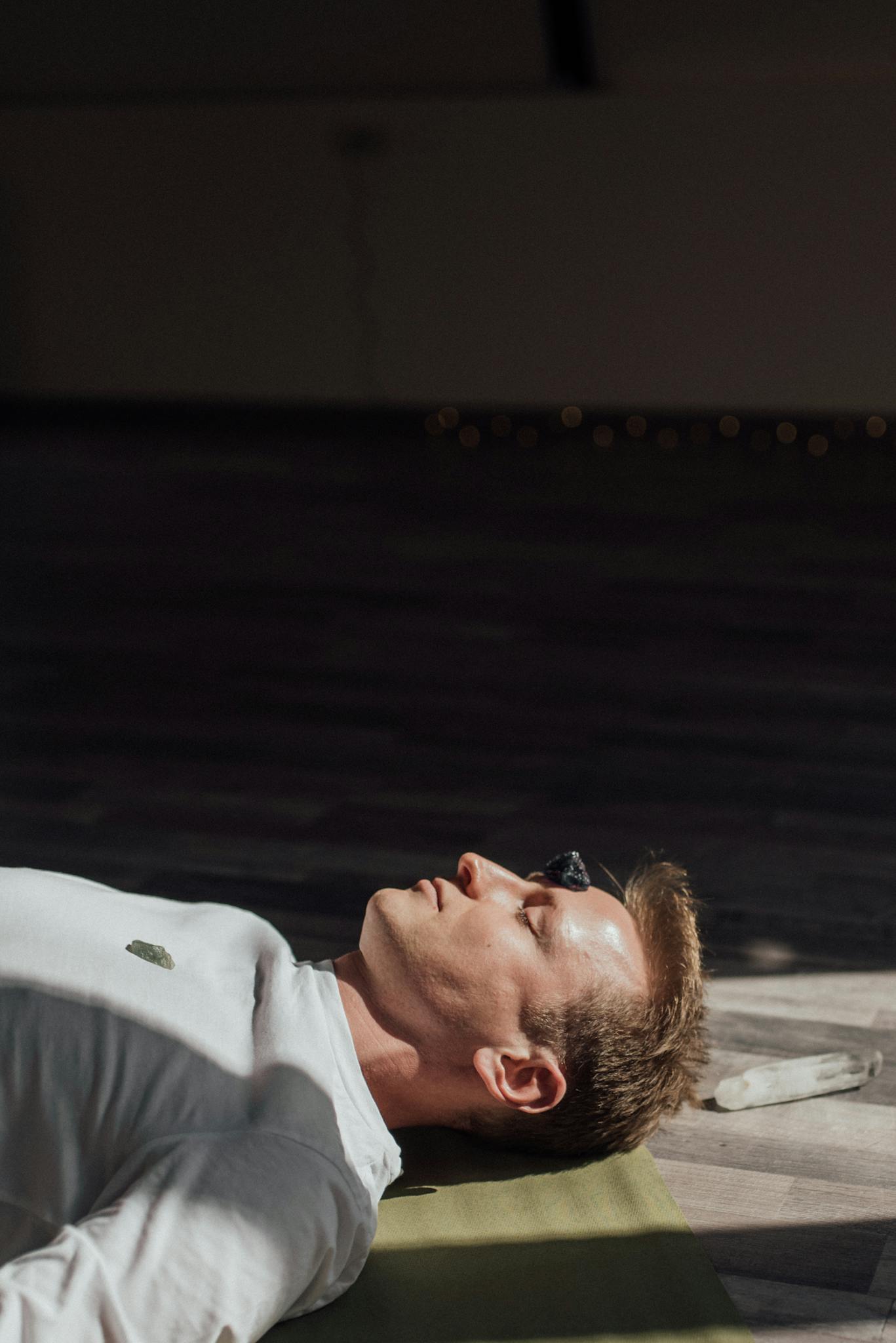 Man meditating indoors with healing crystals on chest and forehead for therapeutic relaxation.